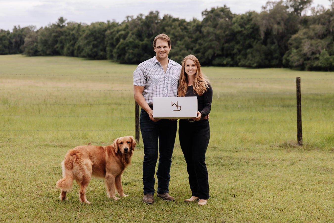 Man and woman holding a laptop with a logo in a grassy field with a dog.