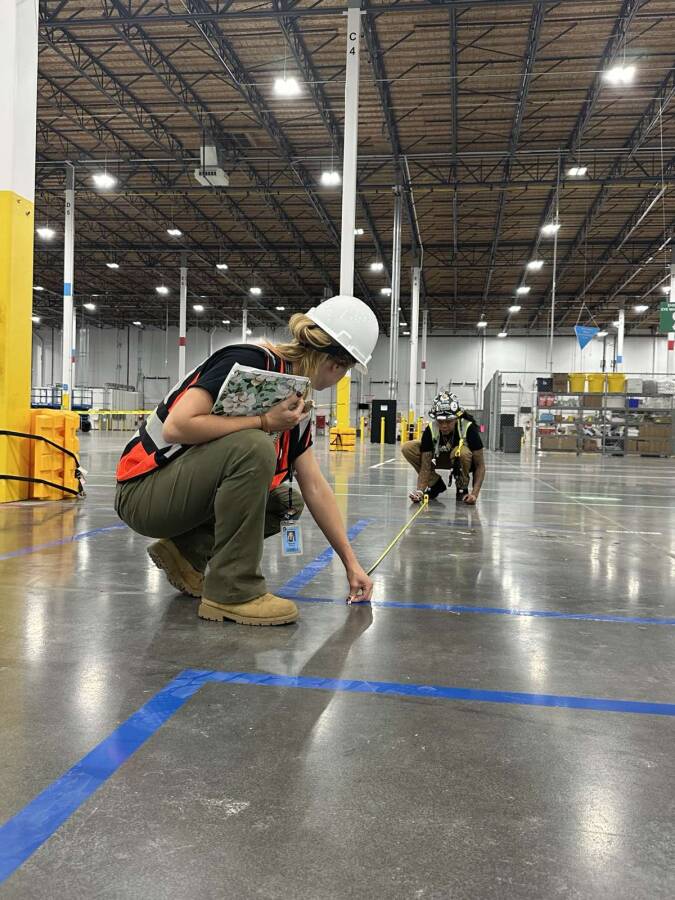 Kelsey kneeling down to in a warehouse floor holding the end of a tape measure while holding a clipboard, wearing hard hat and vest while a co-worker holds the other end of the measure wearing the same. 