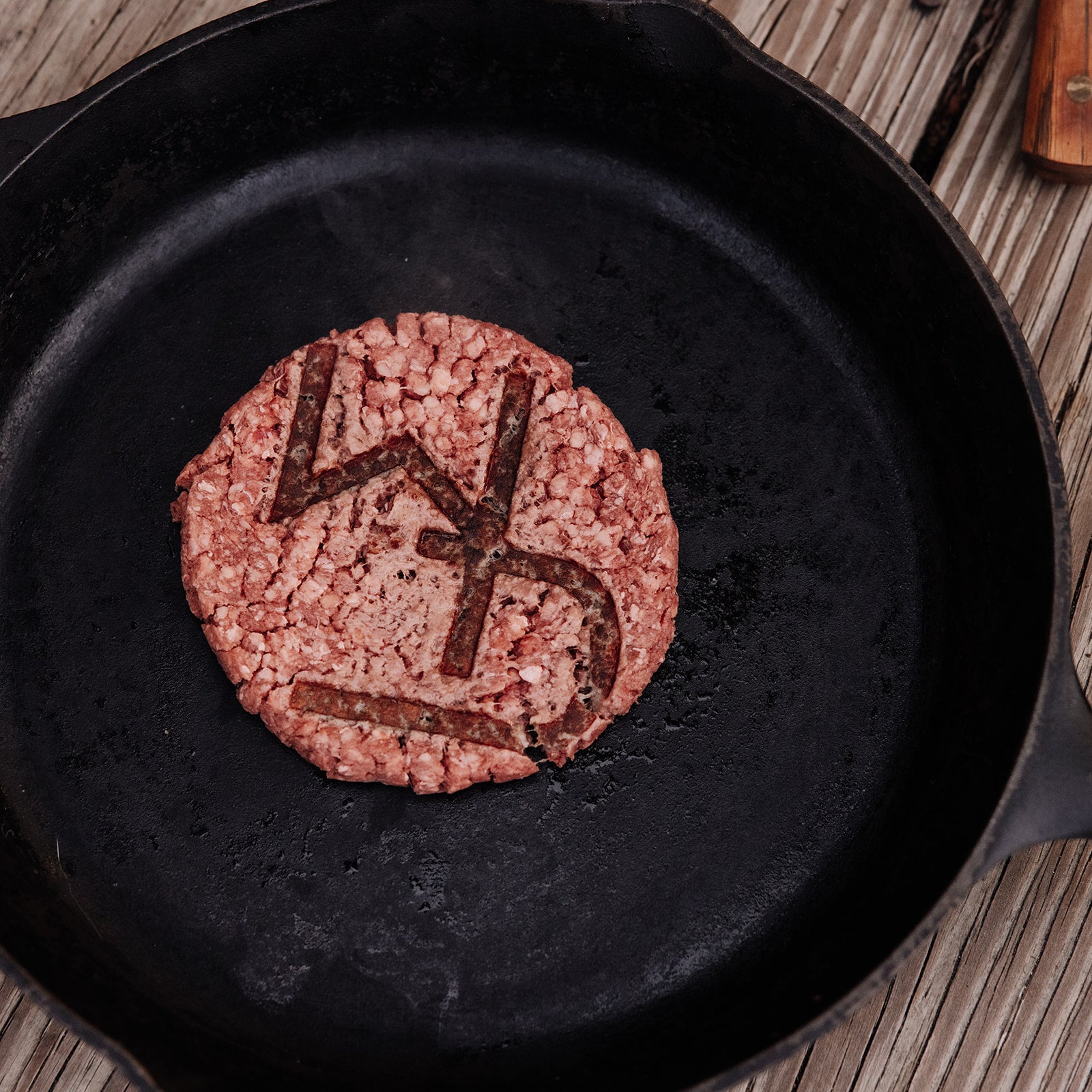 Photography a raw burger patty with the letters "LD" deeply imprinted on its surface, resting in a dark seasoned cast iron pan on a rustic wooden table with a faint knife handle