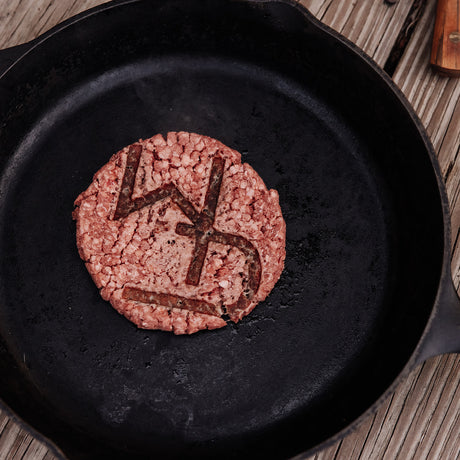 Photography a raw burger patty with the letters "LD" deeply imprinted on its surface, resting in a dark seasoned cast iron pan on a rustic wooden table with a faint knife handle