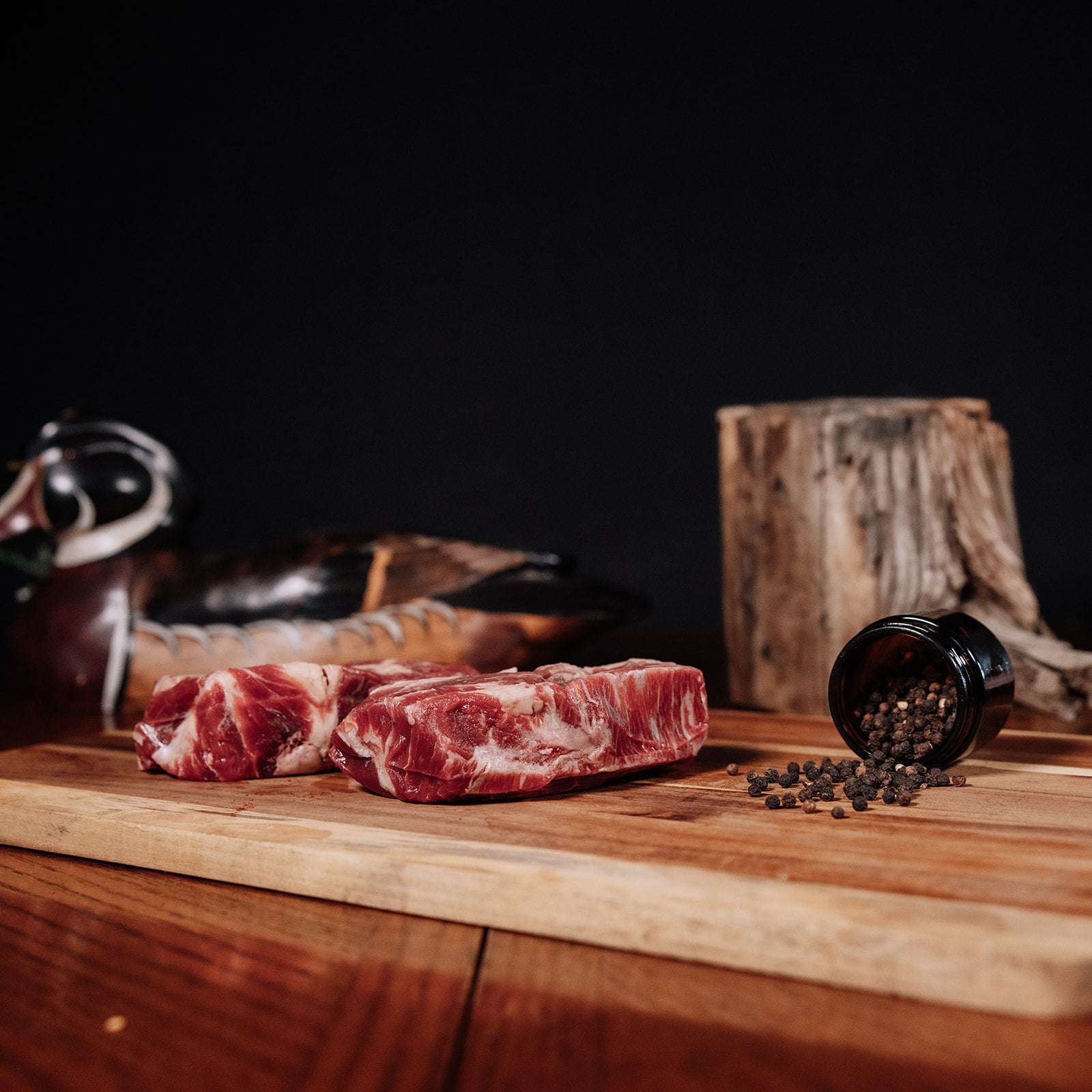 Photograph depicting two marbled raw chuck steaks on a rustic wooden cutting board, accompanied by spilled black peppercorns from a small dark jar, a carved wooden duck decoy, and a weathered tree stump in the background
