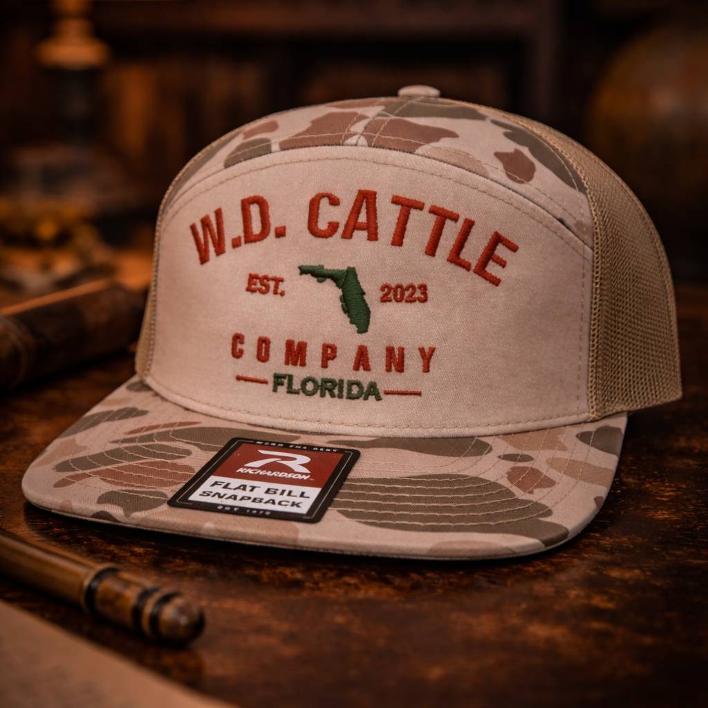Camouflage and beige cap with 'W.D. Cattle Company' branding on a wooden surface.