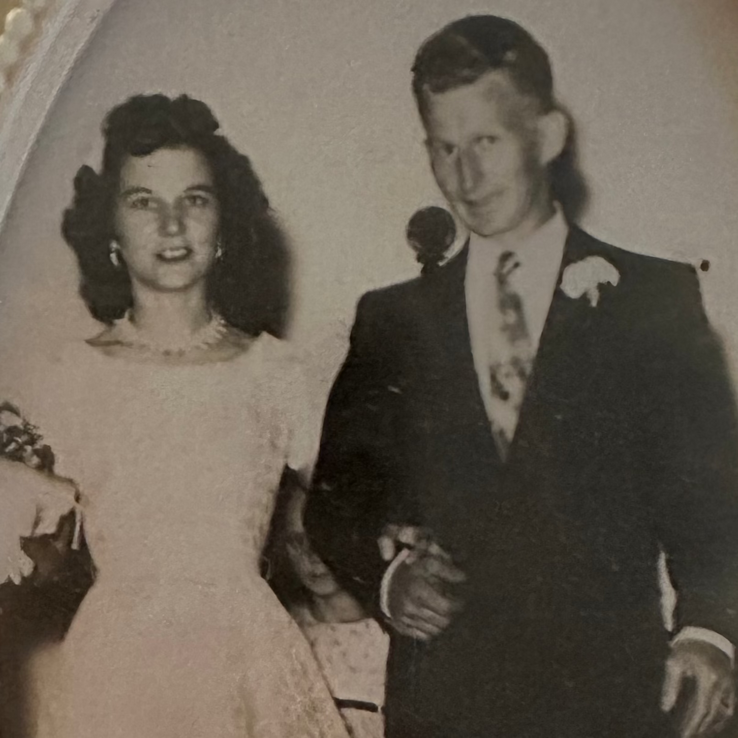 Vintage black and white photo of a couple in their wedding attire - the woman with dark brown and shoulder length hair in a short sleeved wedding dress and necklace next to a young man with a black suit and flower in his lapel. 