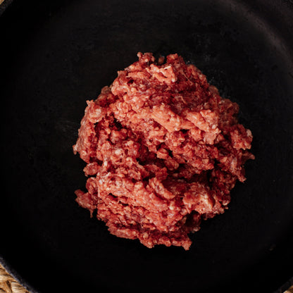 Photograph of raw ground beef in a dark cast iron pan showing bright red and pink colors with visible fat flecks and a coarse texture