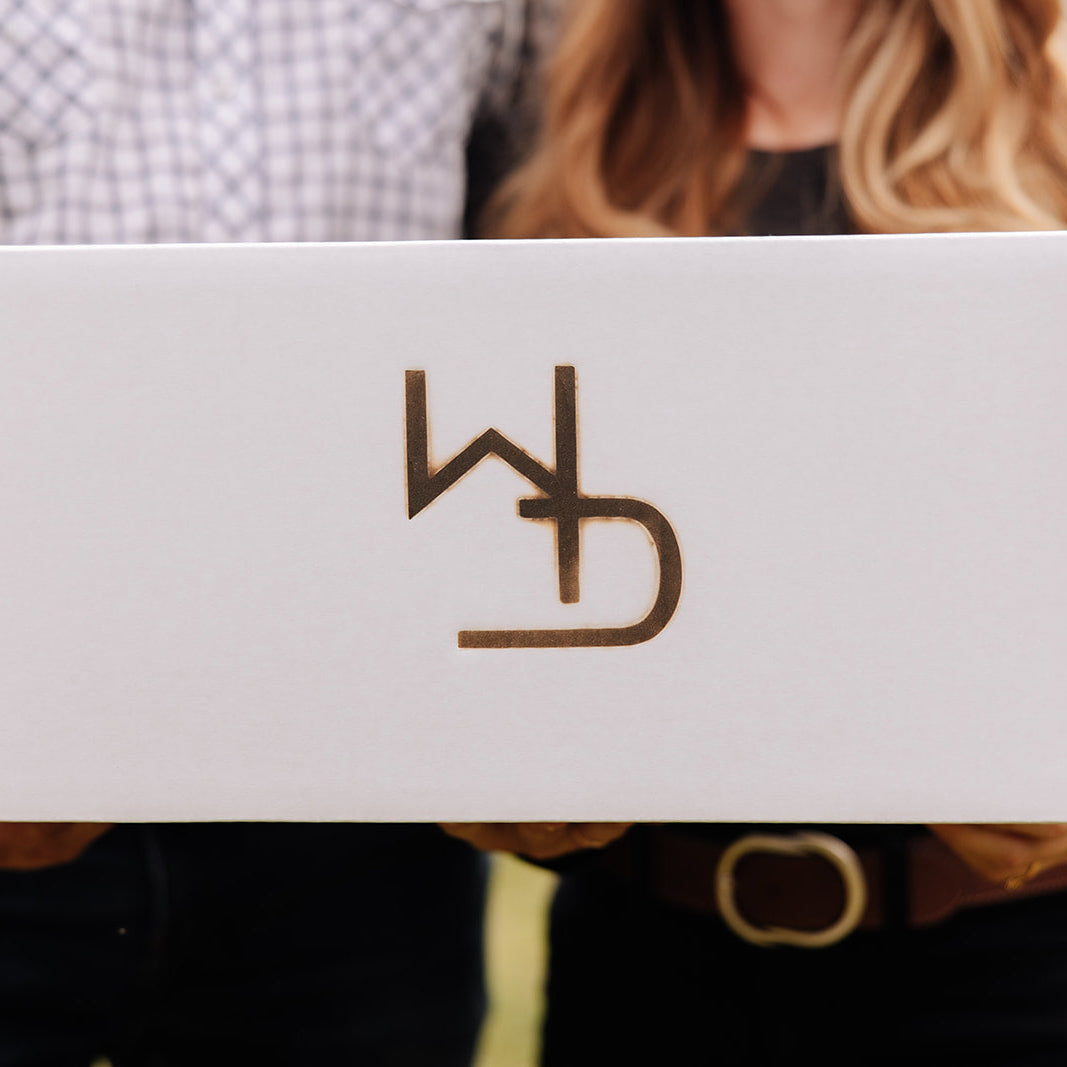 Two people holding a white box with the WD Cattle Company logo branded into the box. Blurred background of two people holding the box together. Brand is a W and a D intertwined on the box.