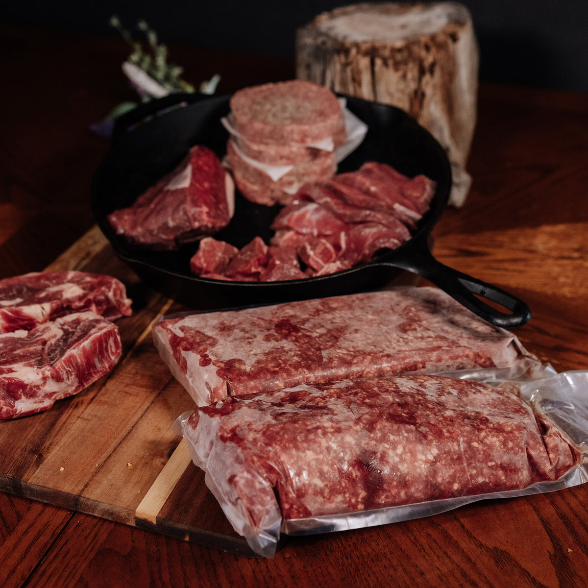 Various raw beef cuts including steaks, ground beef patties in a cast iron skillet, and vacuum-sealed packages of ground meat arranged on a dark wooden table with a cutting board