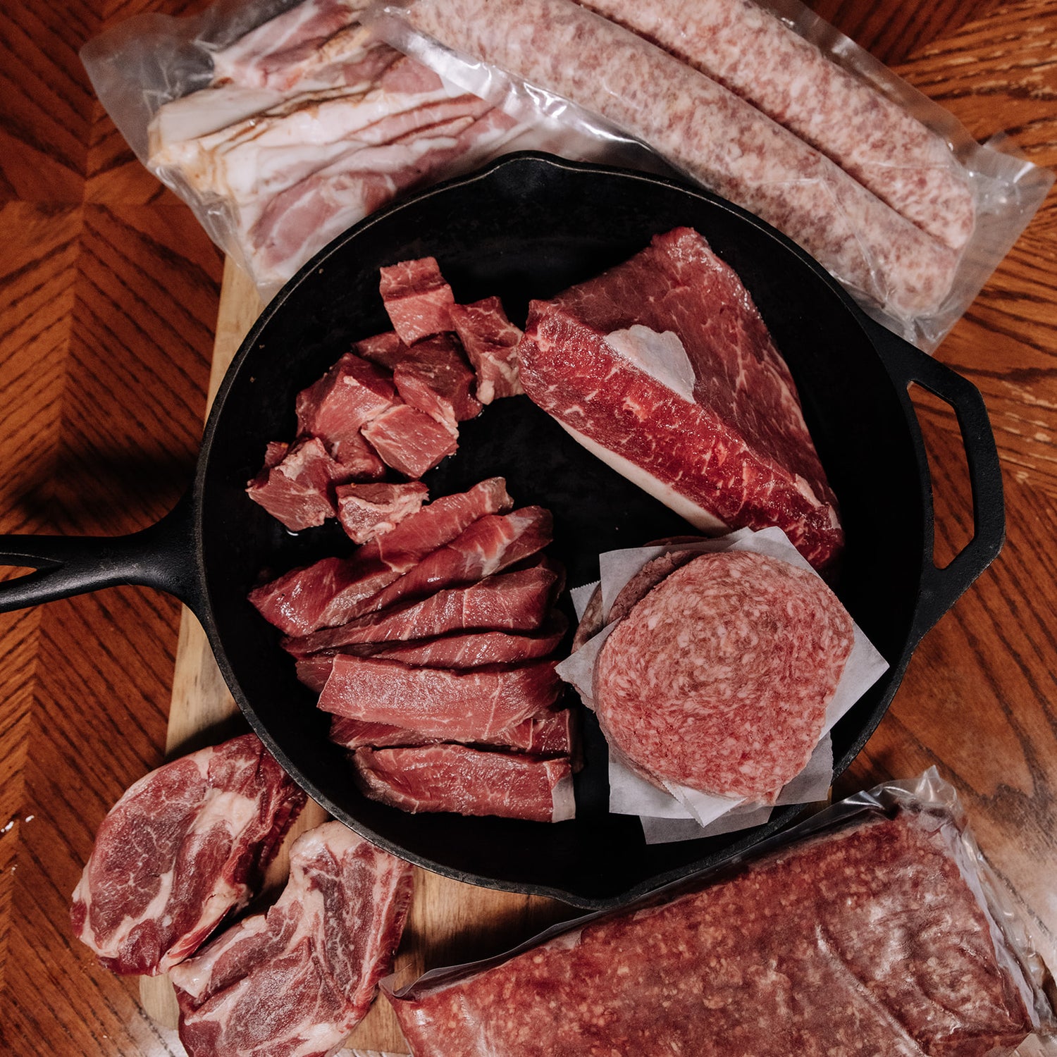 An assortment of fresh raw meats featuring beef cubes, steak cuts, and hamburger patties in a black cast iron skillet, surrounded by vacuum-sealed packages of bacon, sausages, and ground beef on a warm-toned herringbone wood table