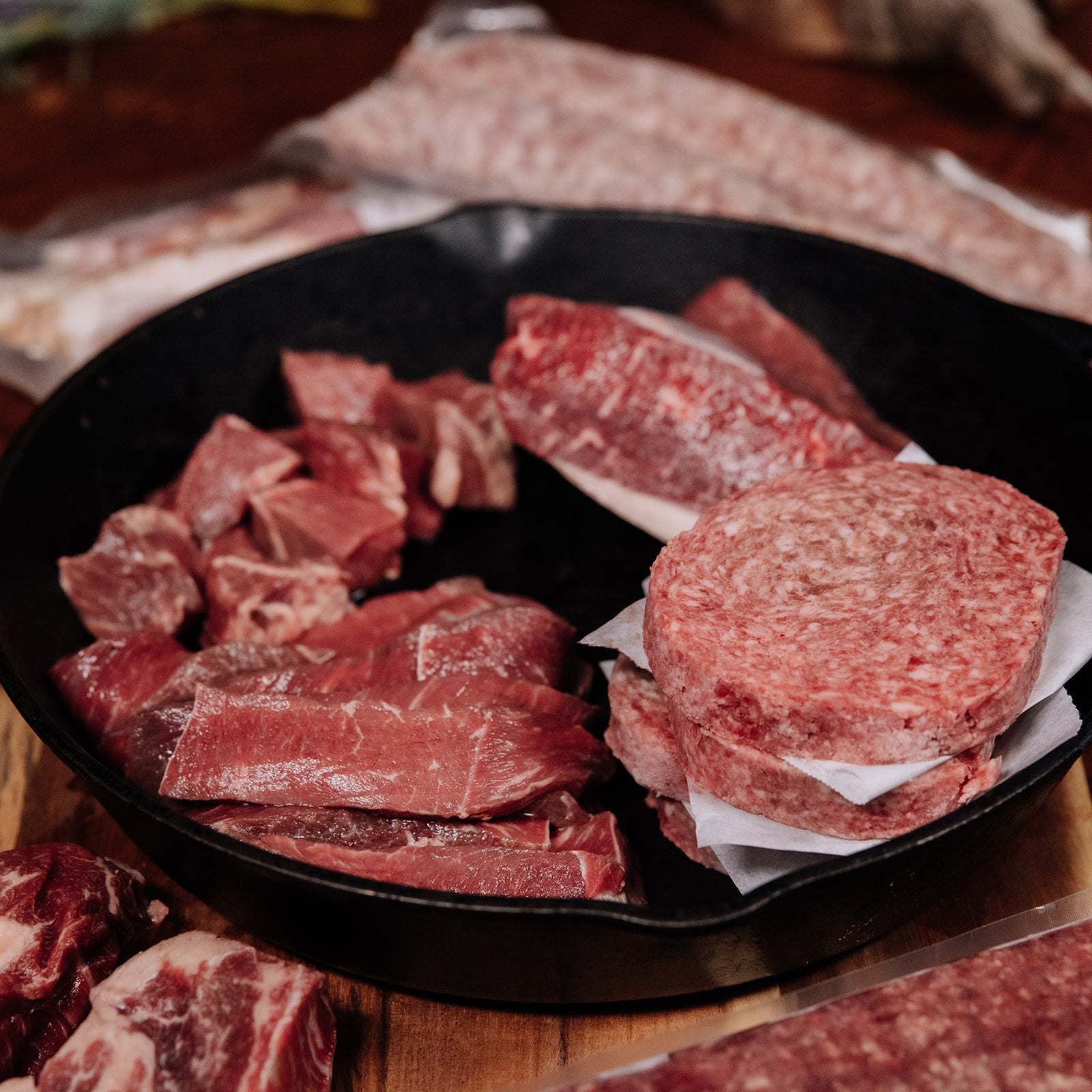 Close-up of a black cast iron pan filled with various raw red meat cuts including stacked burger patties separated by white paper, flat steaks, and cubed beef, arranged on a wooden surface with blurred vacuum-sealed meat packages in the background