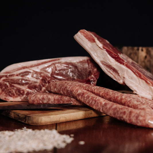 A close-up photograph featuring raw pork shoulder, a fatty slab of pork belly, and several raw Italian sausages on a wooden cutting board with a chef's knife and coarse salt in the foreground against a dark, dramatic background