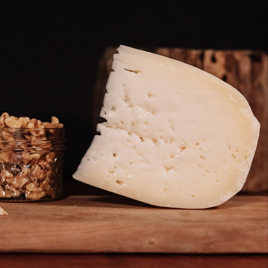 Two types of cheese on a wooden board with a small container of nuts against a dark background