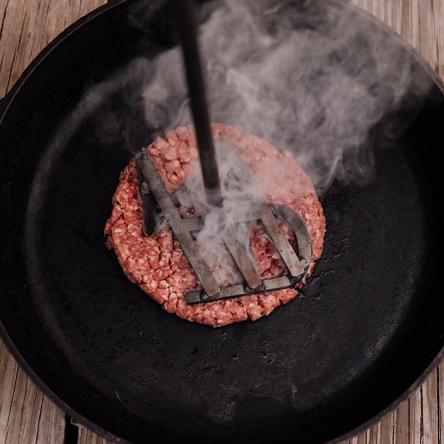 Sizzling raw ground beef patty in a dark cast iron pan as a metal branding iron is pressed into it, emitting white steam on a rustic wooden surface