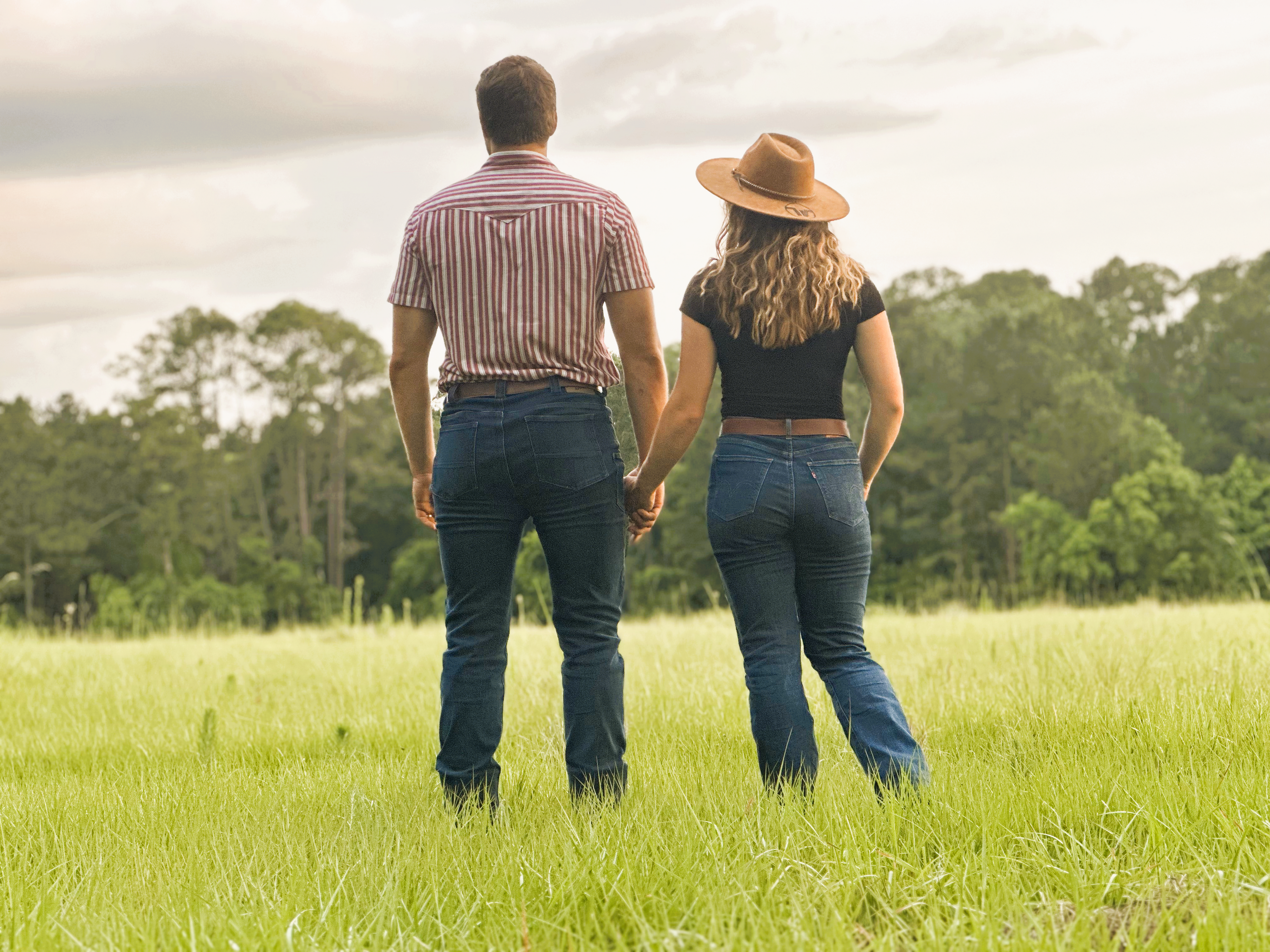 Kelsey and Tyler - a man and woman - holding hands in a grassy field with trees in the background and their backs to the camera. 