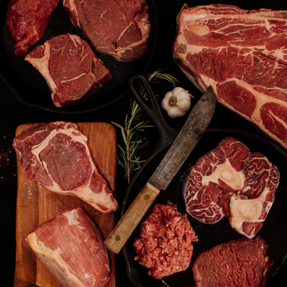 Overhead still life of raw beef cuts featuring ribeye steaks, osso buco, and ground beef on dark cast iron cookware and a wooden board, accompanied by a rustic cleaver, fresh rosemary, garlic, and sea salt flakes