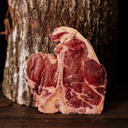 A raw T-bone steak rests on a dark wooden table against a textured tree trunk showcasing rich red marbling and a thick white bone