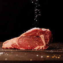 Photography a raw, richly marbled ribeye steak on a dark wooden surface with coarse salt falling onto its vibrant red flesh against a stark black background