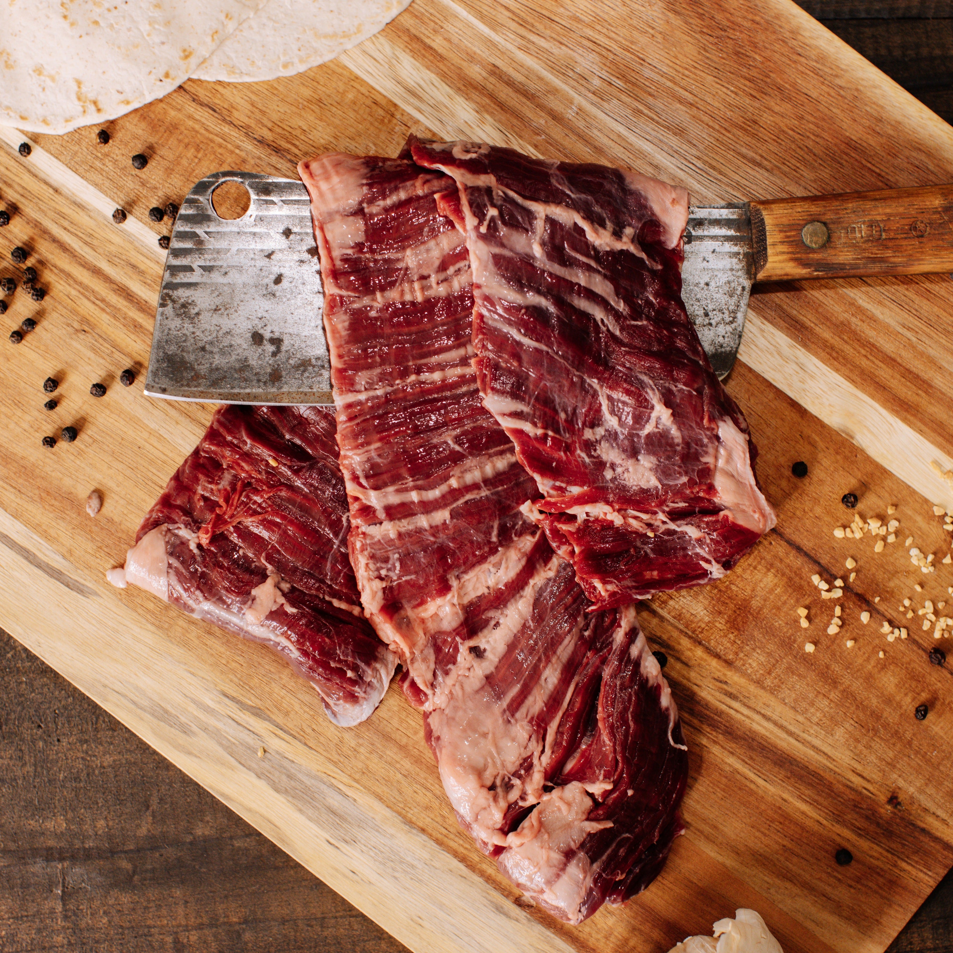 Photograph of raw dark red skirt steak with white marbling on a striped wooden cutting board, alongside a rustic meat cleaver with a wooden handle, scattered black peppercorns, and white tortillas