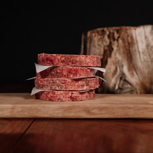Photograph of four raw burger patties stacked on a light wooden cutting board, each separated by white parchment paper, displaying vibrant red and white marbled meat textures against a dark background with a blurred rustic wooden log