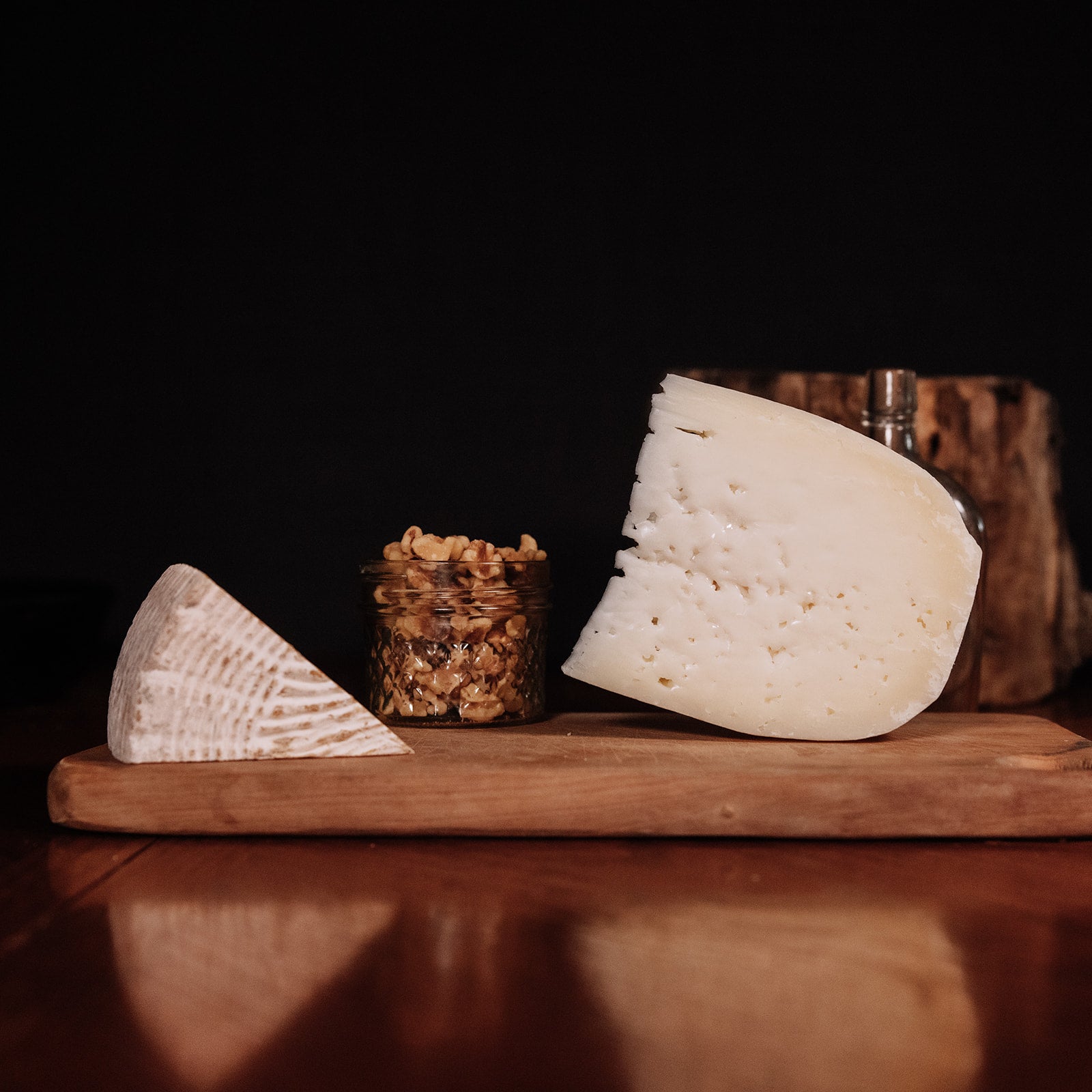 Wedge of swiss cheese standing on a cutting board next to a jar of walnuts and Tomme cheese wedge lying on its side with a dark background