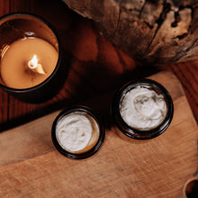 Photography of a warm glowing candle in a black holder beside two open amber jars of white whipped tallow lotion on a rustic wooden board 