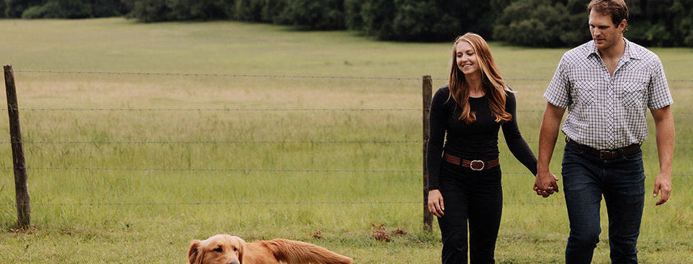 Two people walking a dog in a grassy field with trees in the background