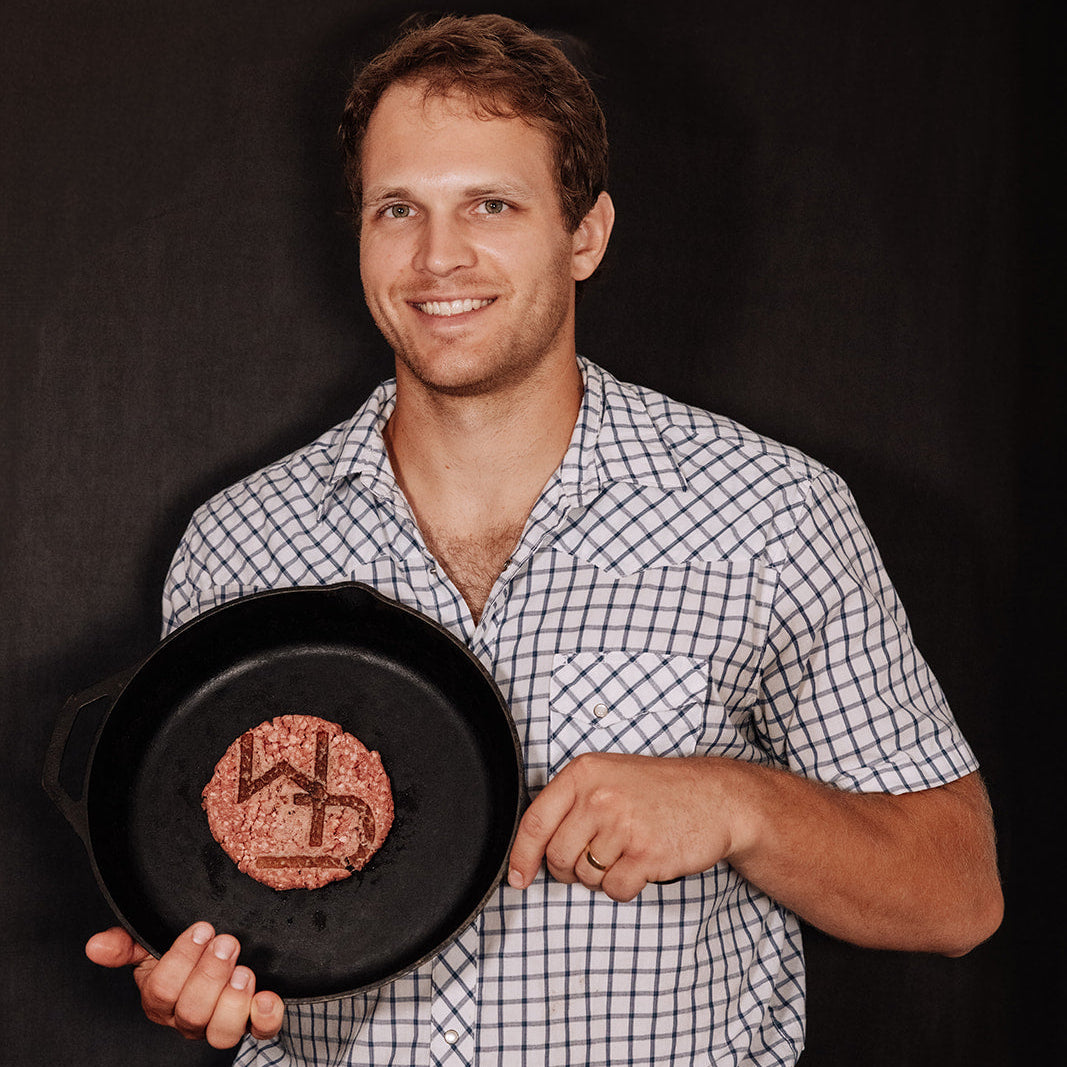 Tyler smiling and holding a skillet with a burger patty in it. 