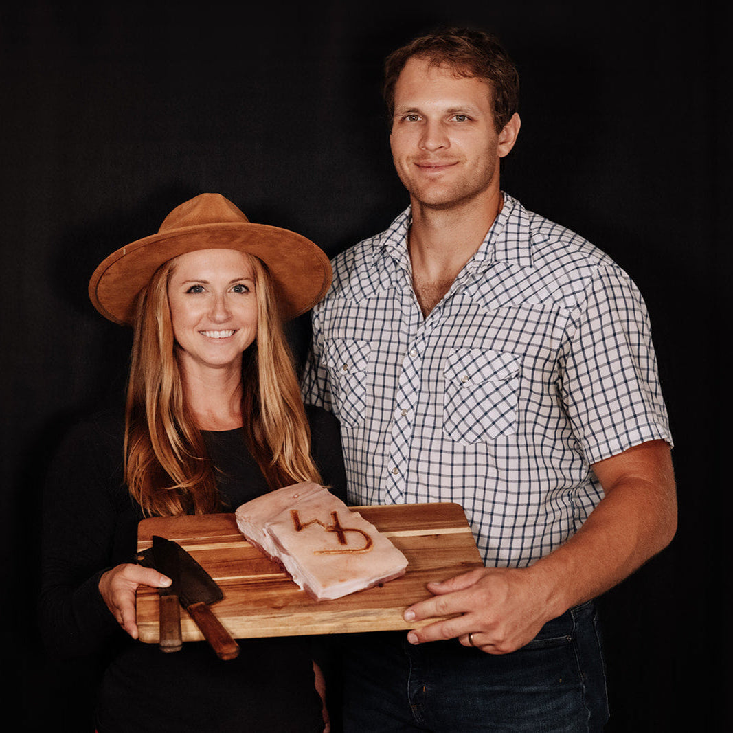 WD Cattle Company owners - Kelsey and Tyler, standing in front of a black background, smiling and holding a cutting board with a piece of pork belly and a butchers knife resting on the cutting board. The WD Cattle Company logo branded into the pork belly. Brand is a W and a D intertwined.