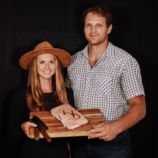 WD Cattle Company owners - Kelsey and Tyler, standing in front of a black background, smiling and holding a cutting board with a piece of pork belly and a butchers knife resting on the cutting board. The WD Cattle Company logo branded into the pork belly. Brand is a W and a D intertwined.