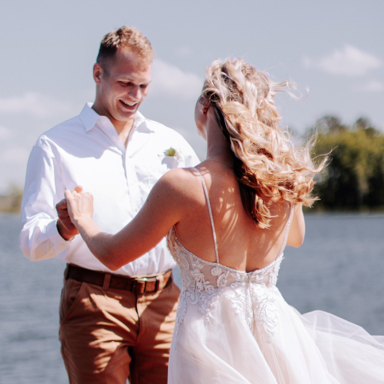 Kelsey and Tyler standing on a dock with the wind blowing Kelseys hair and dress and Tyler smiling as they have their first look event. A body of water with trees in are blurred in the background