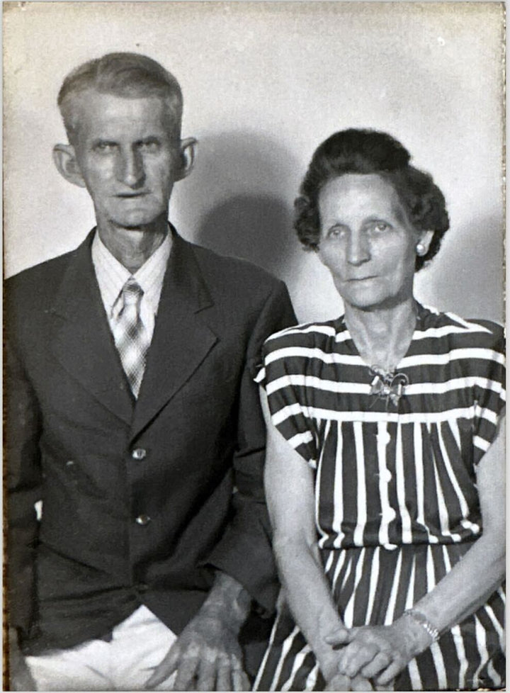 Black and white photo of an elderly couple sitting together, the lady wearing a striped dress and the man a suit. 
