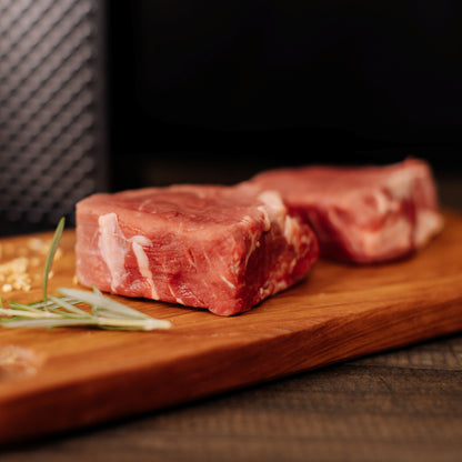 Photography of two raw red meat steaks with white marbling on a wooden cutting board, garnished with fresh green rosemary sprigs and golden chopped garlic against a dark textured background