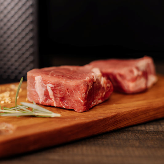 Photography of two raw red meat steaks with white marbling on a wooden cutting board, garnished with fresh green rosemary sprigs and golden chopped garlic against a dark textured background