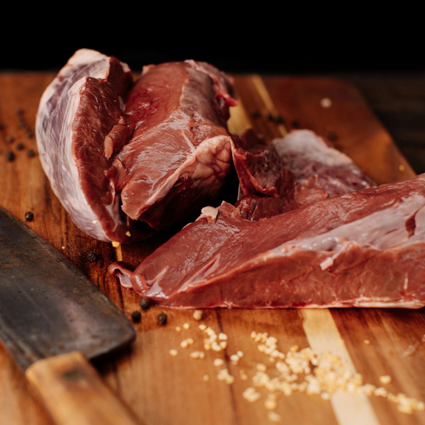 Fresh dark red meat portions on a warm wooden cutting board beside a rustic butcher cleaver and scattered black peppercorns and white garlic
