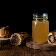 A clear glass jar filled with golden yellow liquid stands on a wooden board, surrounded by several raw bone pieces on a dark rustic tabletop against a black background.